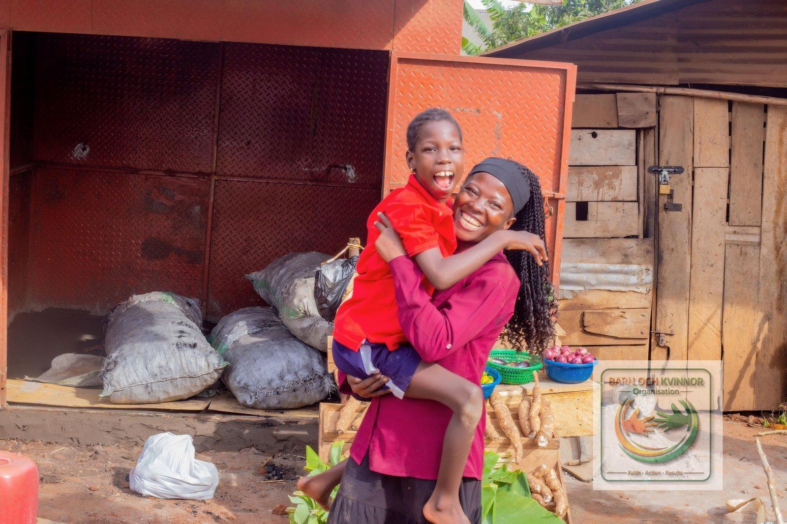 Mama Jennifer holding her smiling daughter in front of her empty kiosk